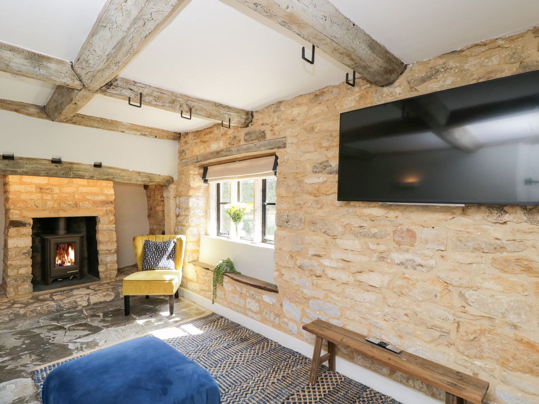 A living room with a stone fireplace yellow chair window with flowers blue ottoman and wall mounted television at Millbank Cottage in Mickleton
