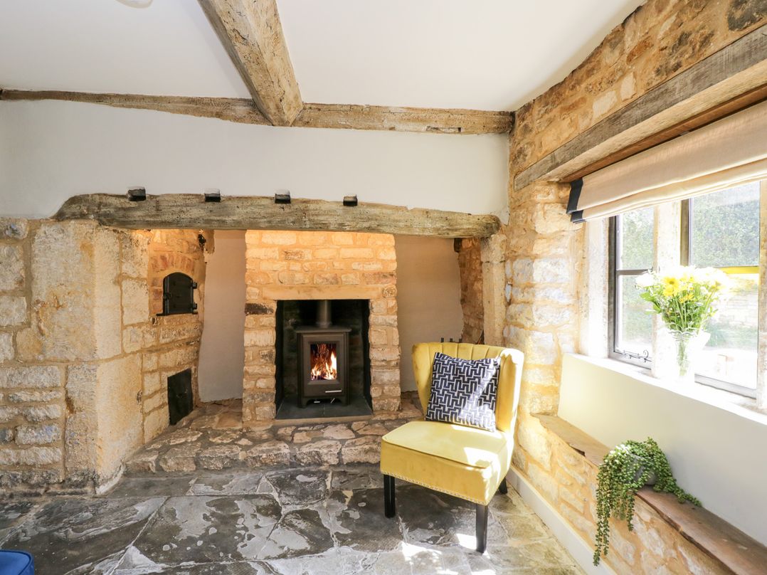 A room with a stone fireplace a yellow chair with a cushion and flowers on the window sill at Millbank Cottage in Mickleton
