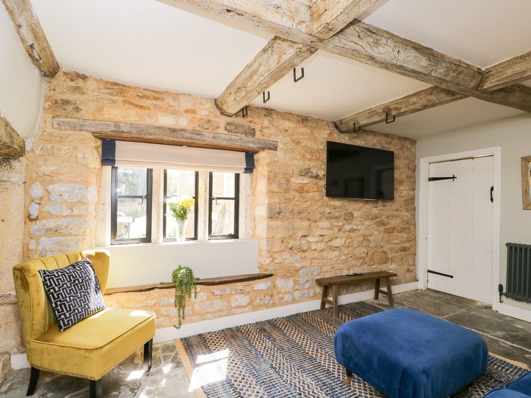 A room with stone walls a yellow chair a blue ottoman a wall-mounted tv and wooden ceiling beams at Millbank Cottage in Mickleton
