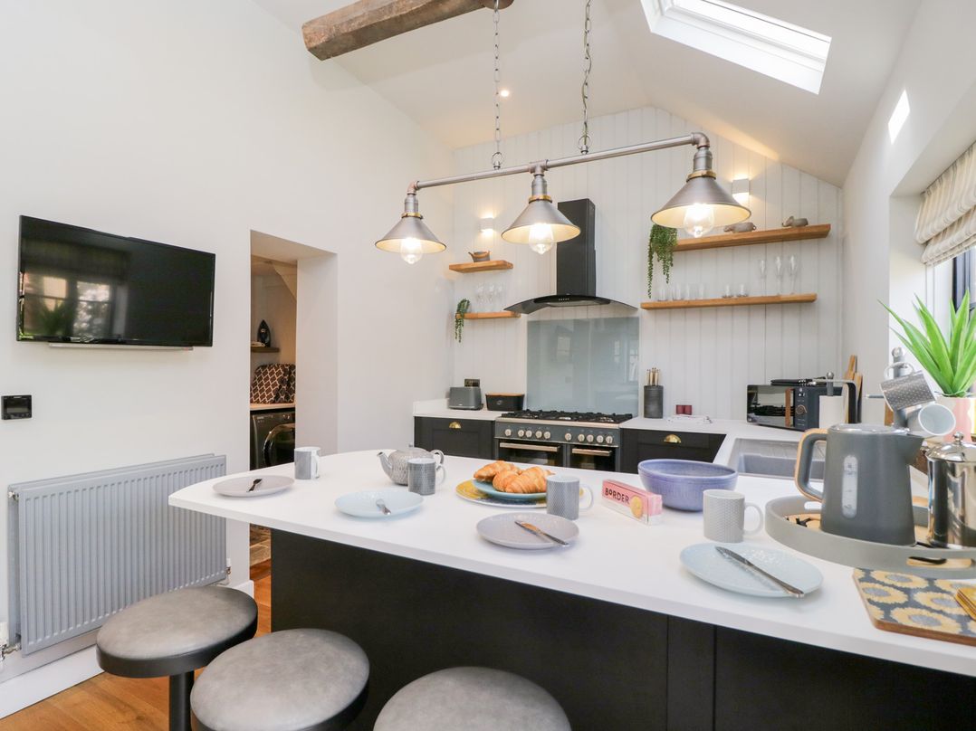 A kitchen with a white island counter set with plates mugs and croissants a black stove and hood wooden shelves and a skylight at Millbank Cottage in Mickleton