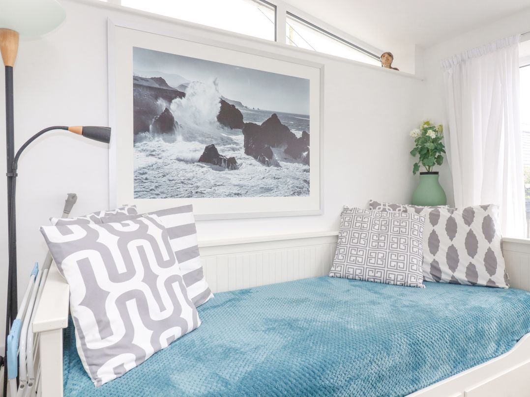 A white daybed with patterned cushions and a blue blanket next to a wall with a large framed ocean wave photo and a green vase with flowers at Langurra in Crantock