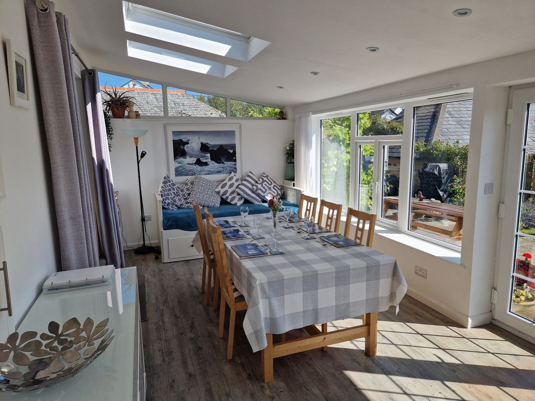 A dining room with a long table and wooden chairs next to large windows and a daybed with patterned cushions at Langurra in Crantock