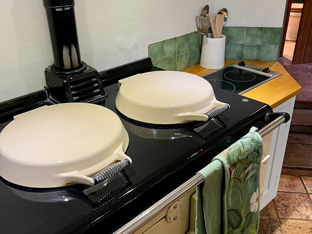 A kitchen with a black stove with cream covers a ceramic hob and kitchen utensils in a white container at Meadow View Cottage in Calcot near Northleach
