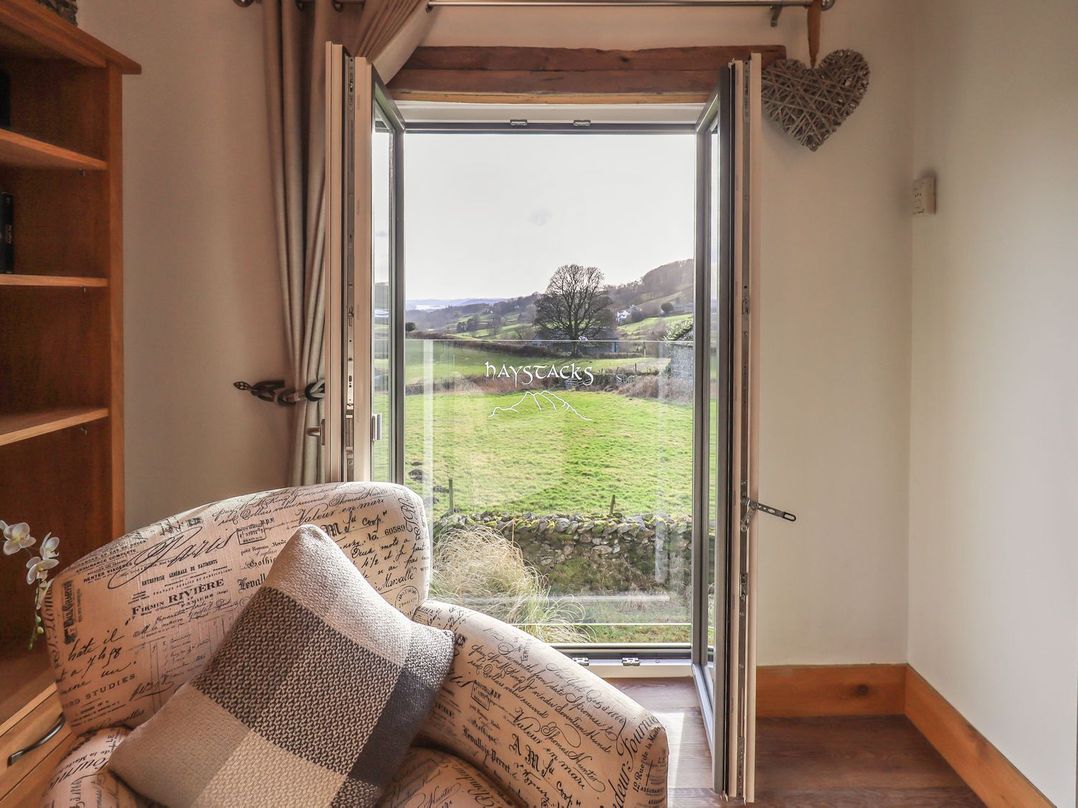 A room with a patterned armchair and a pillow facing an open glass door showing a green field at Haystacks in Troutbeck