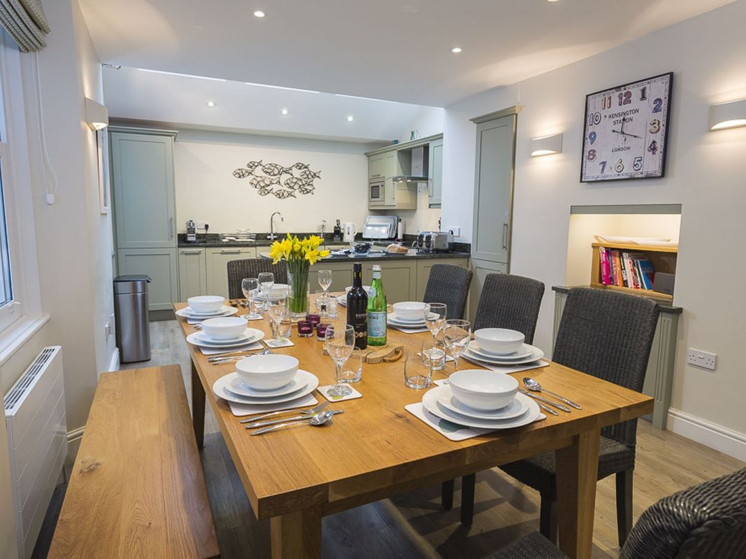 A dining area with a wooden table set with plates glasses cutlery and a vase of flowers next to a kitchen with green cabinets at Leylands in Salcombe