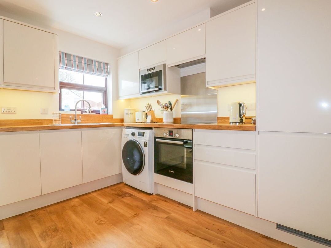 A kitchen with white cabinets wooden countertops a washing machine oven microwave kettle and a window with a striped blind at The Beach Hut in Crantock near Newquay