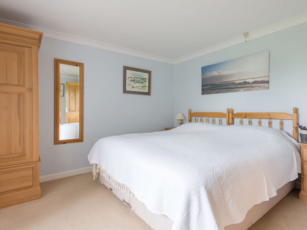 A bedroom with a double bed covered in a white bedspread a wooden wardrobe mirror and a nightstand lamp at Curlew in Rock