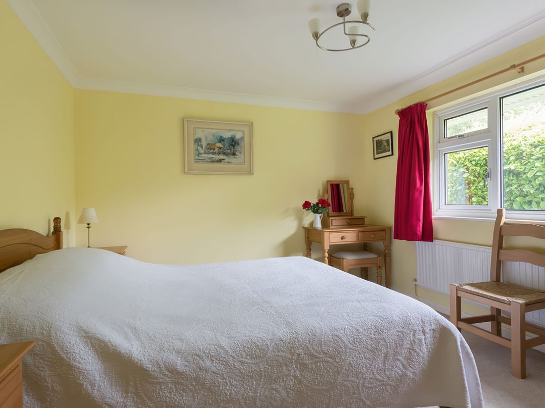 A bedroom with a bed covered in a white quilt a wooden dressing table with flowers a window with red curtains and a wooden chair at Curlew in Rock