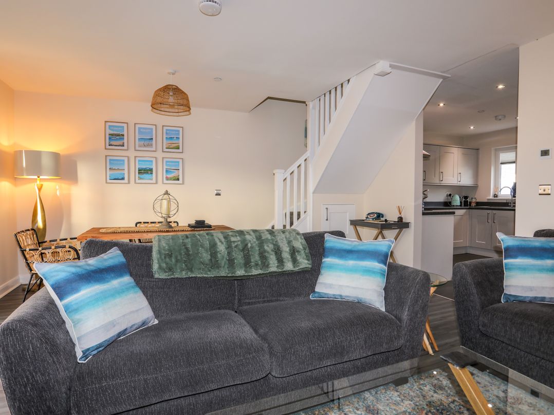 A living room with gray sofas with blue pillows a dining table with chairs wall art and a kitchen in the background at 19 Parc Delfryn in Benllech