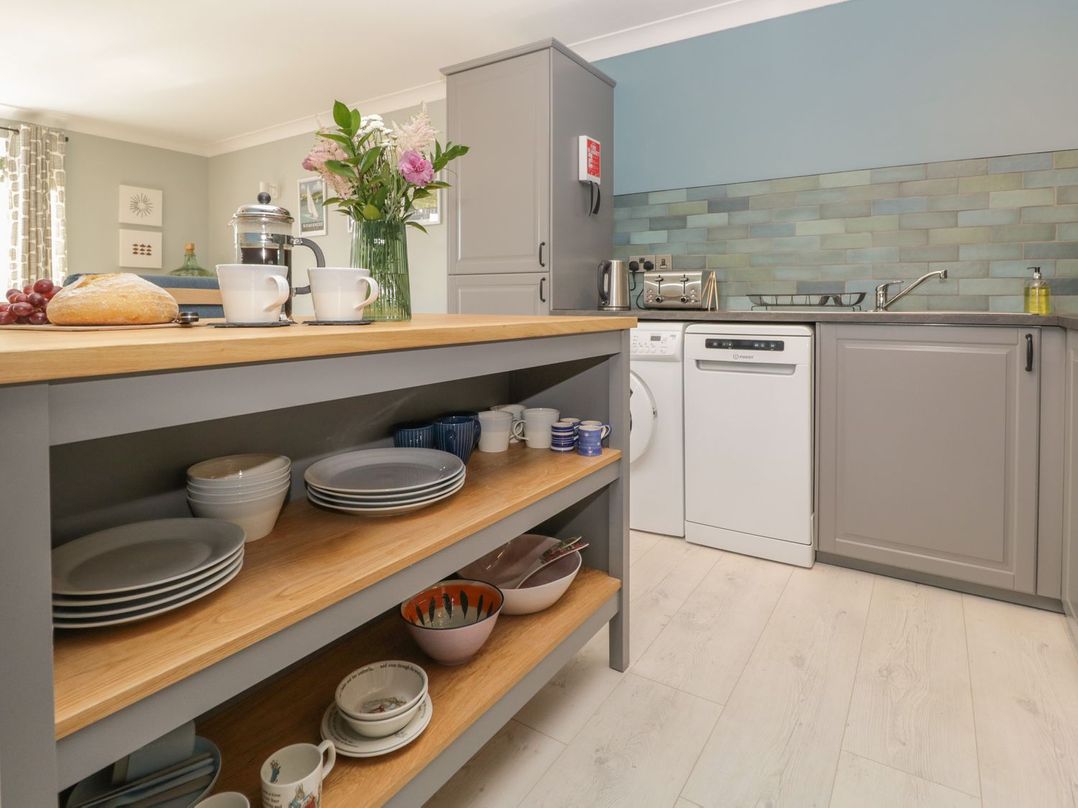A kitchen with grey cabinets a wooden countertop a kitchen island with shelves holding dishes mugs and bowls and a vase of flowers at Newlands in Windermere