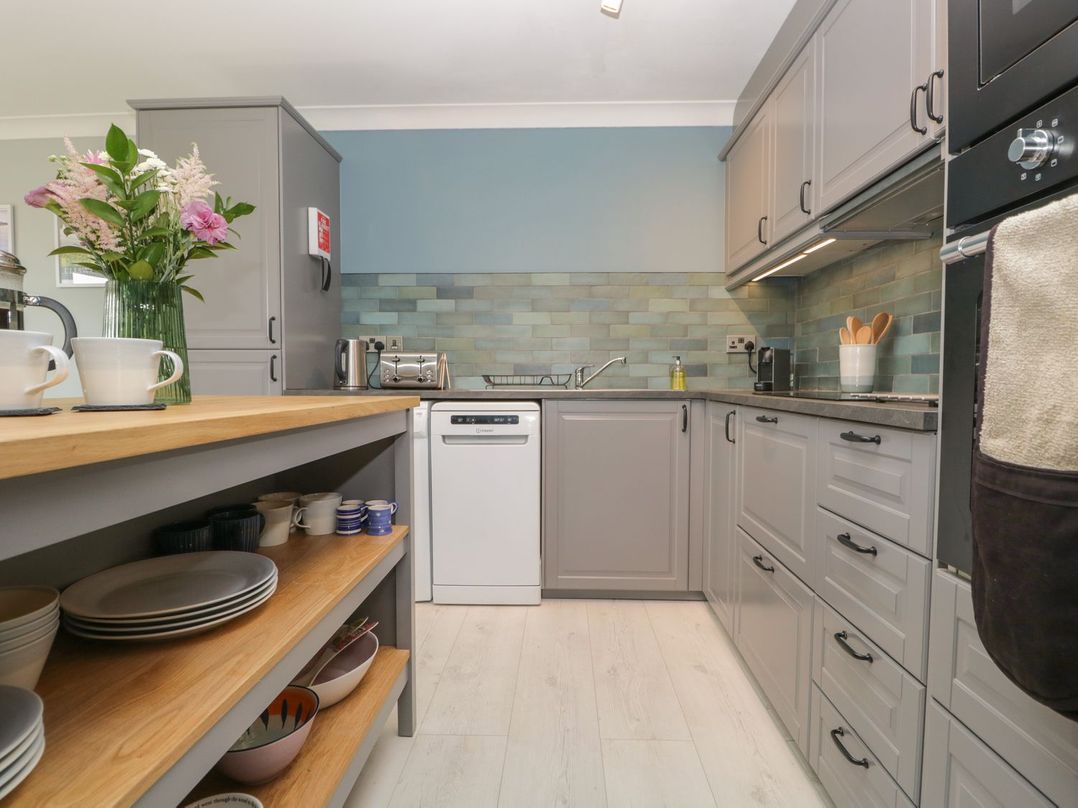 A kitchen with grey cabinets a wooden countertop open shelves with dishes and mugs and a dishwasher at Newlands Windermere