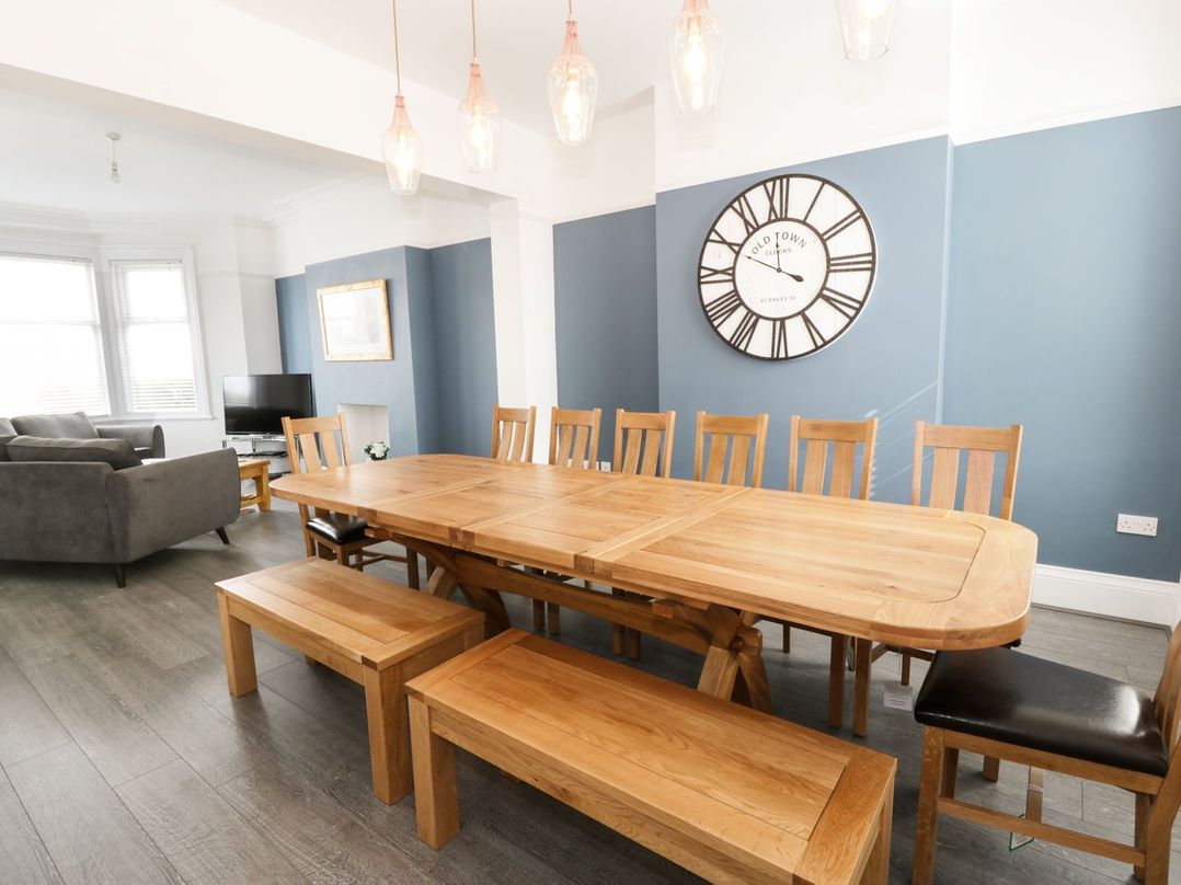 A dining room with a large wooden table surrounded by chairs and benches and a living area with sofas and a television at 51 Trinity Avenue in Llandudno