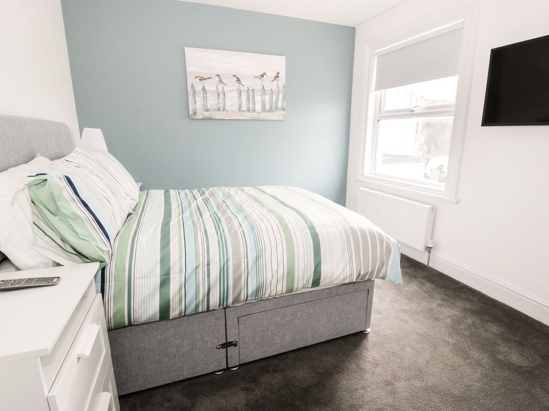 A bedroom with a bed with striped bedding a bedside table a lamp a window with a blind and a wall-mounted television at 51 Trinity Avenue in Llandudno