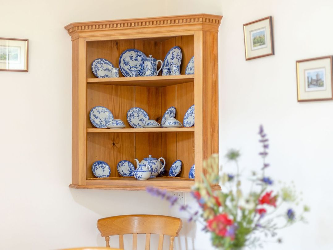 A wooden corner shelf with blue and white china dishes on the wall above a wooden chair with a blurred flower vase in the foreground at Swallow's Nest in Strefford near Craven Arms