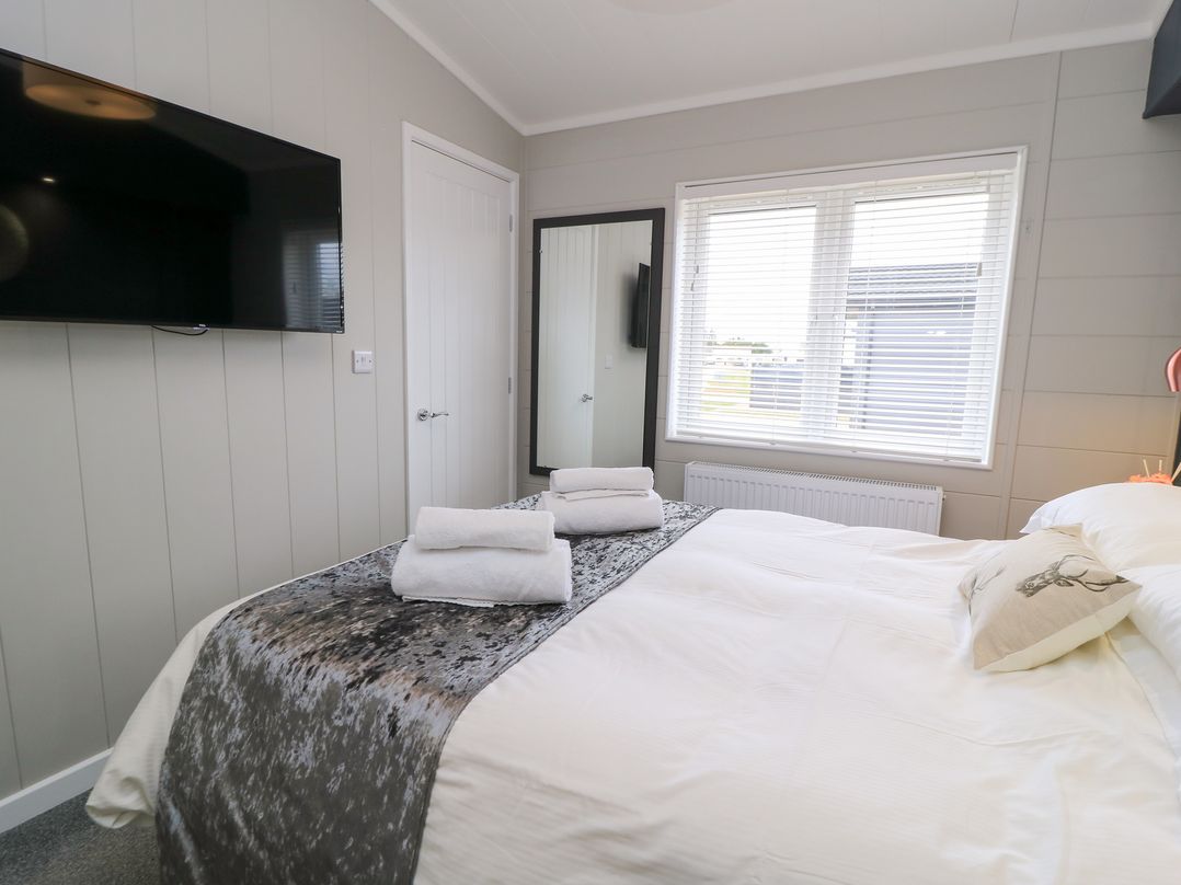 A bedroom with a bed with white linen and folded towels a wall mounted tv a window with blinds and a mirror at The Gallops in Chipping Norton