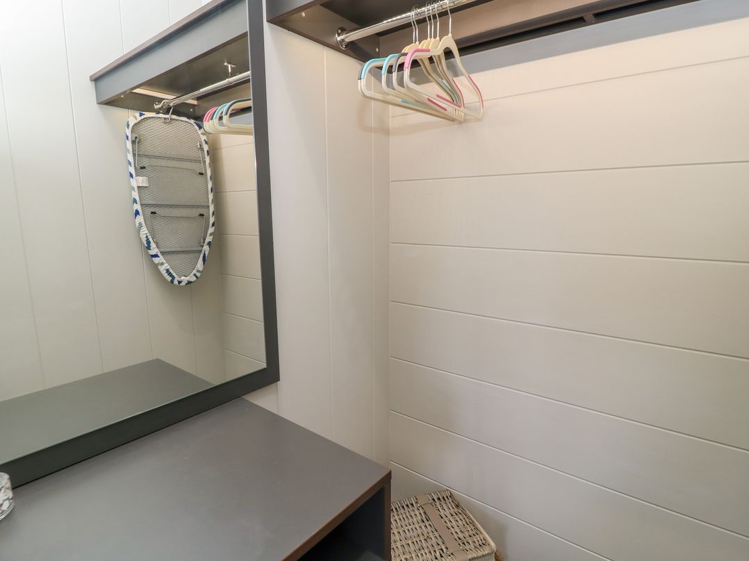 A small room with an empty clothes rail with hangers a mirror an ironing board and a woven basket at The Gallops in Chipping Norton