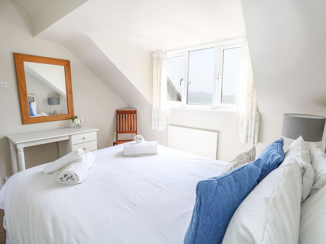 A bedroom with a bed with blue pillows white curtains a wooden chair a white desk and a mirror at Sea Spray in Bowleaze