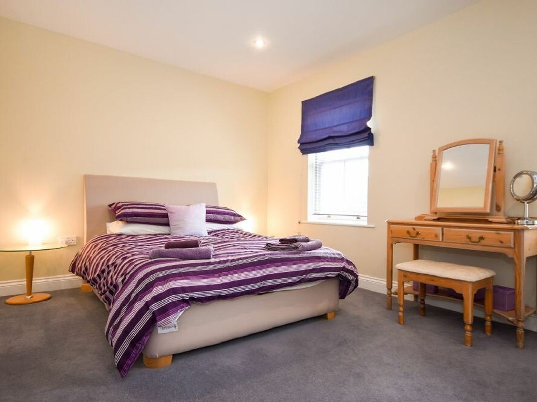 A bedroom with a bed with striped bedding a round side table with a lamp and a wooden dressing table with a mirror and stool at Churchside in Alnwick