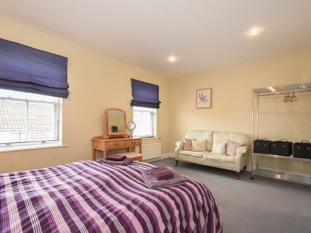 A bedroom with a bed covered in a purple striped blanket a wooden dressing table with a mirror two windows with blue blinds a white sofa and a metal clothes rack at Churchside in Alnwick