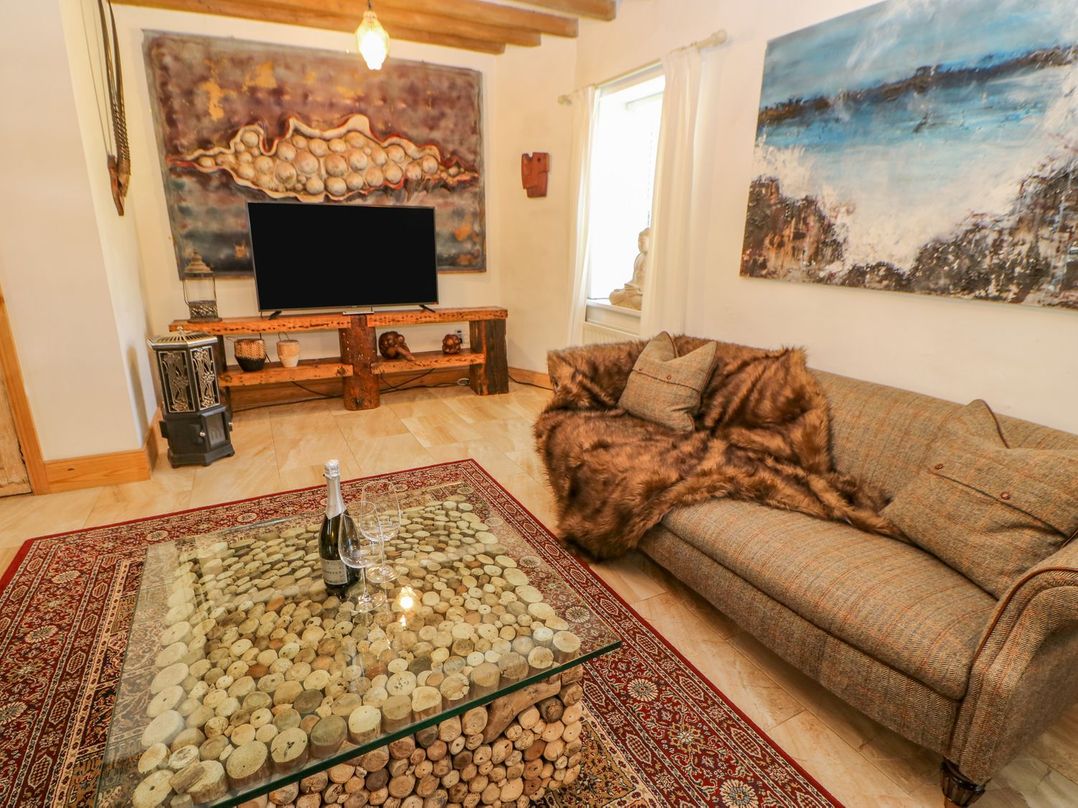 A living room with a sofa with cushions and fur throw near a coffee table with wine bottle and glasses and a TV on wooden stand at Shaftoe Mews in Haydon Bridge