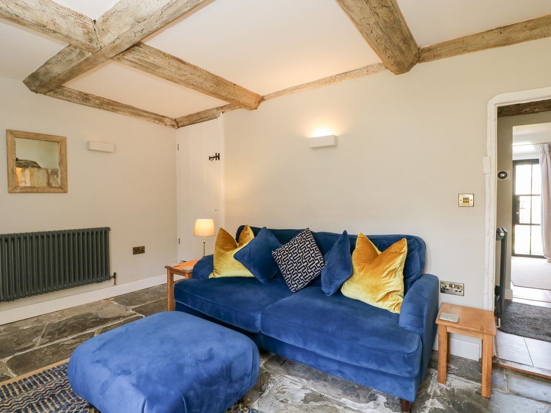 A seating area with a blue sofa and ottoman with yellow and patterned cushions in a room with stone flooring and wooden beams at Millbank Cottage in Mickleton