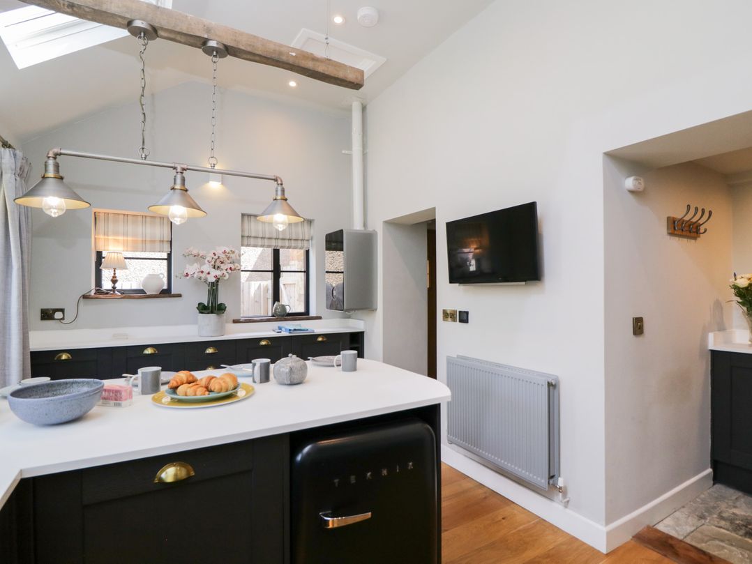A kitchen with an island countertop with croissants and mugs a black fridge under the counter pendant lights windows with shades and a wall-mounted TV at Millbank Cottage in Mickleton