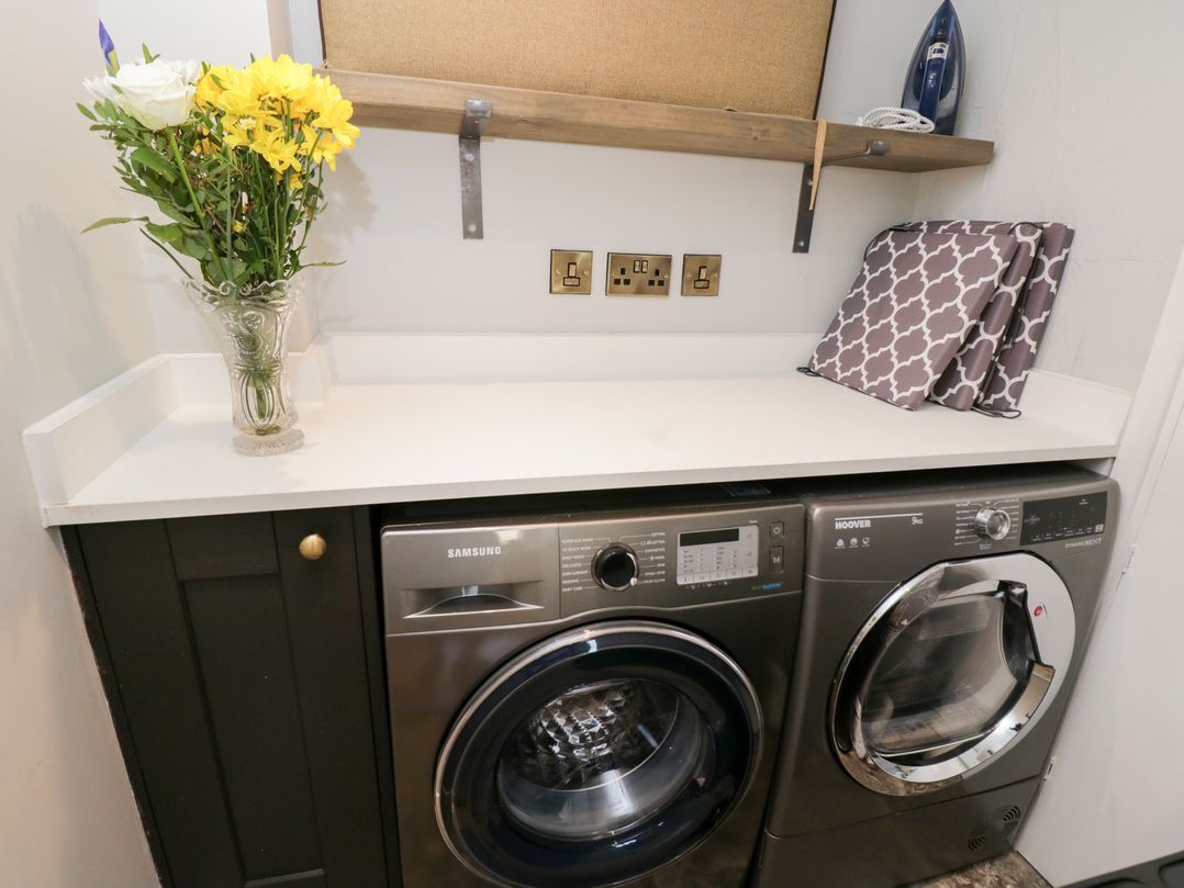 A laundry room with a Samsung washing machine Hoover dryer flowers on a countertop and an iron on a shelf at Millbank Cottage Mickleton