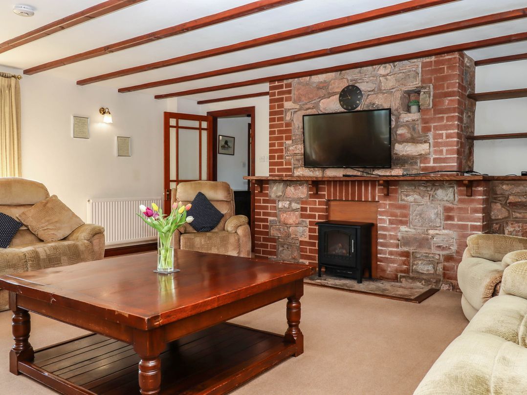 A living room with beige armchairs a wooden coffee table with flowers a mounted television and a brick fireplace at Valley House Halberton near Tiverton