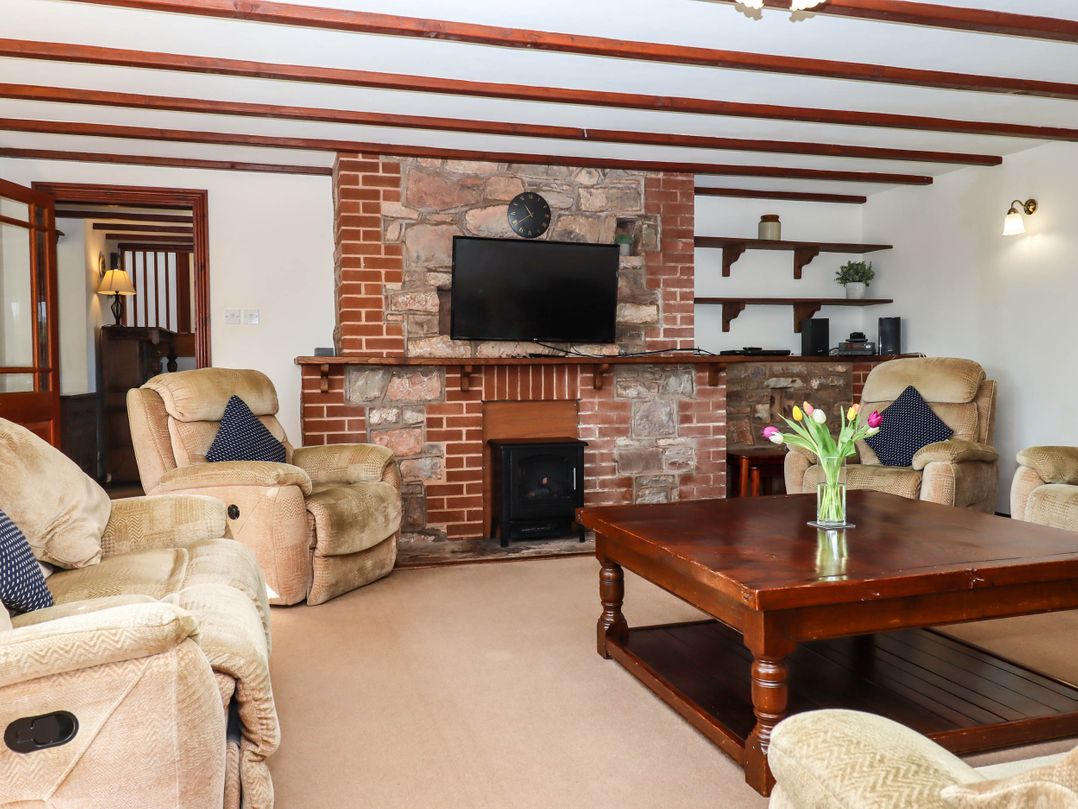 A living room with beige armchairs a wooden coffee table with flowers and a stone fireplace with a mounted TV at Valley House in Halberton near Tiverton