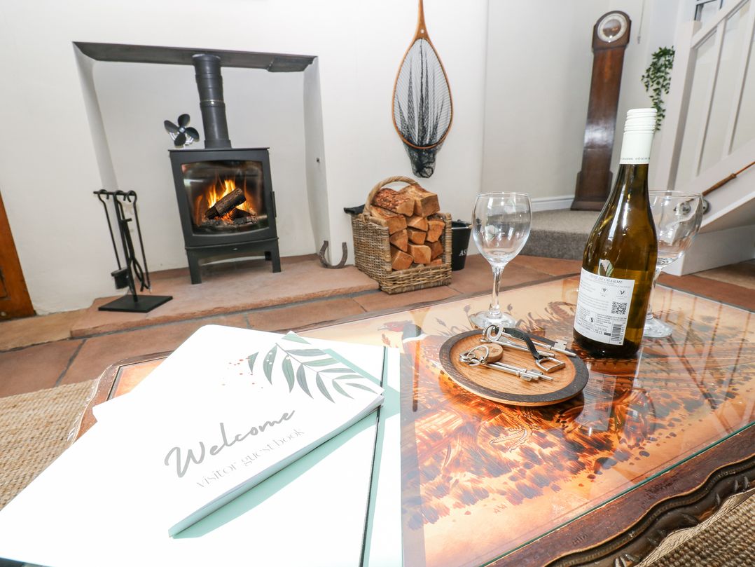 A room with a wood-burning stove fire a basket of firewood a glass table with a bottle of wine wine glasses keys and a welcome visitor guestbook at 3 Eden View in Lazonby