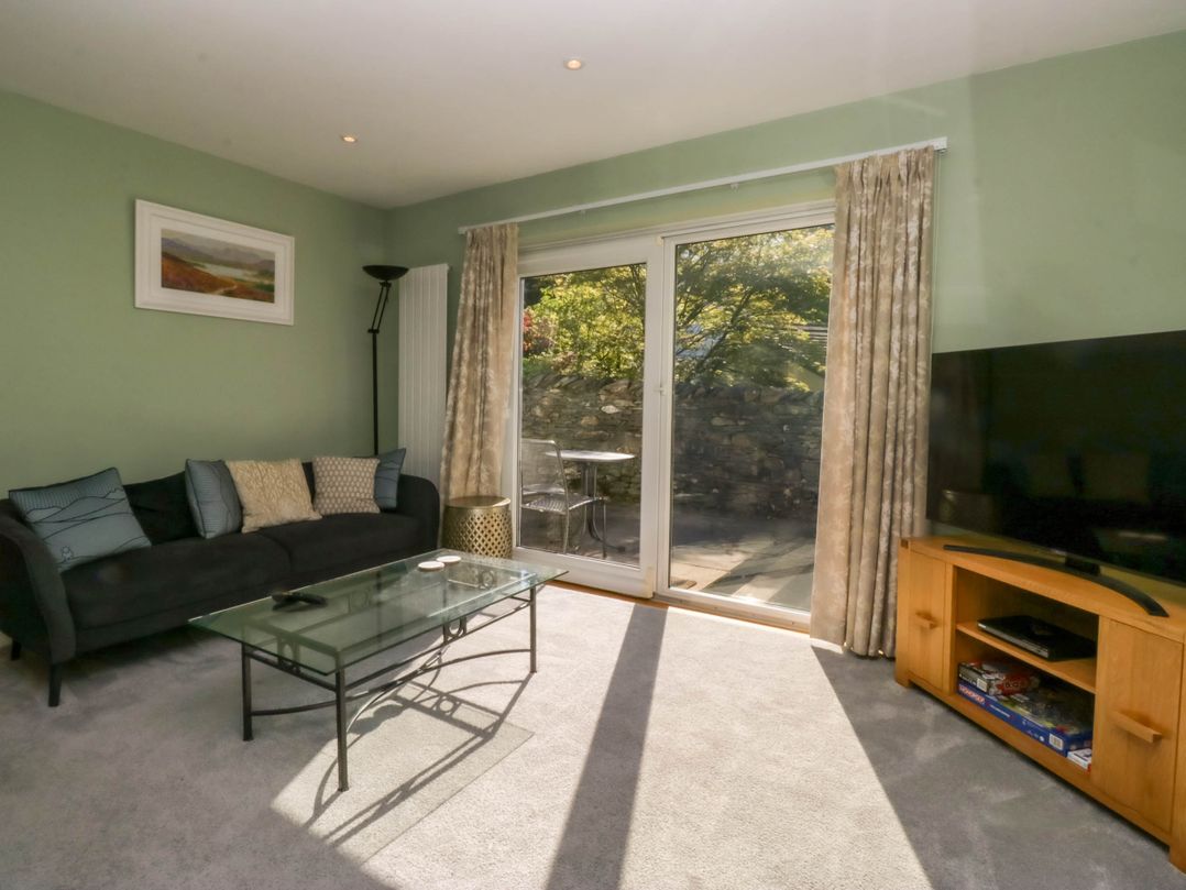 A living room with a black sofa and cushions a glass coffee table a wooden TV stand with a television and patio doors leading outside at Beckside in Ambleside