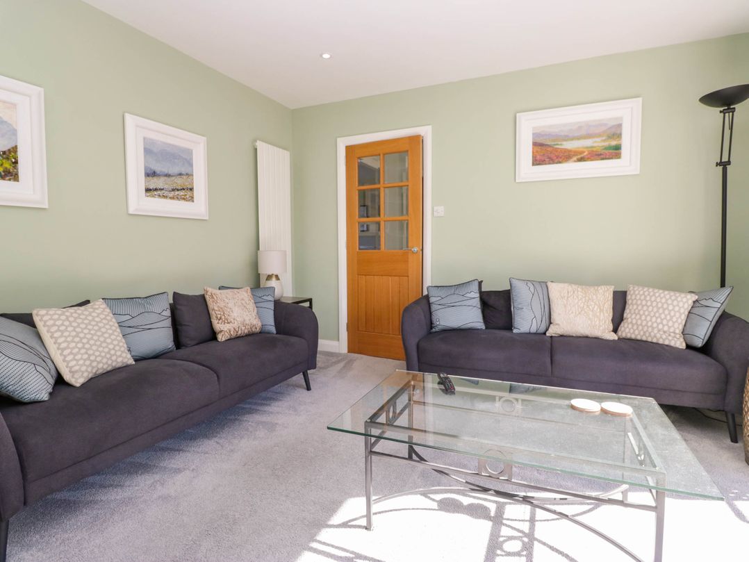 A living room with two dark sofas and multiple cushions a glass coffee table lamp wooden door and framed pictures on green walls at Beckside Ambleside