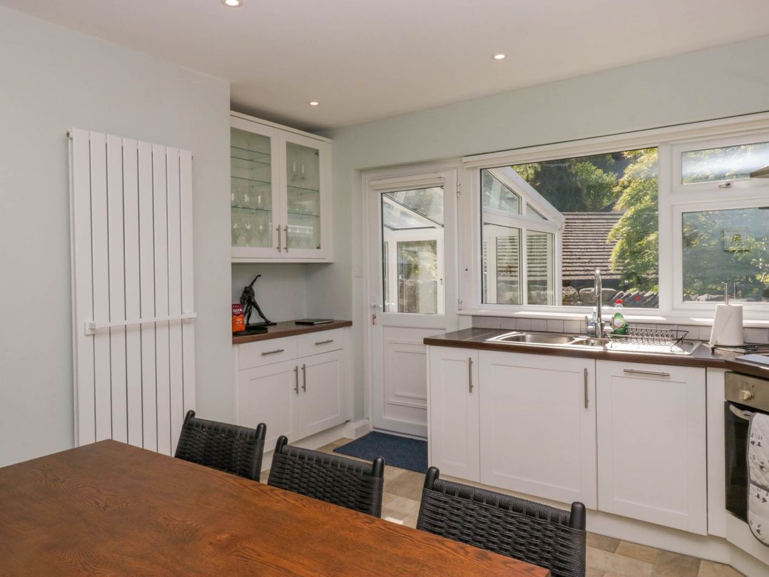 A kitchen with white cabinets a wooden dining table black woven chairs and a window showing greenery at Beckside in Ambleside