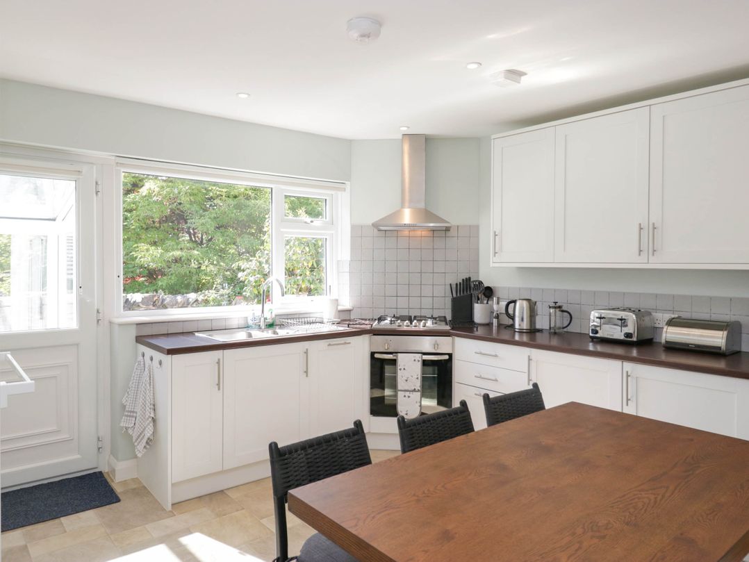 A kitchen with white cabinets wooden countertop dining table and black chairs with a window showing trees at Beckside in Ambleside