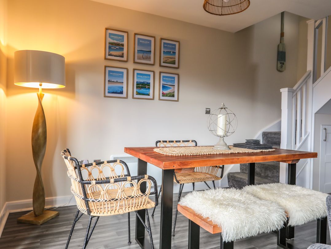 A dining area with a wooden table surrounded by wicker chairs and benches covered with white fur throws a floor lamp and framed pictures on the wall near stairs at 19 Parc Delfryn in Benllech