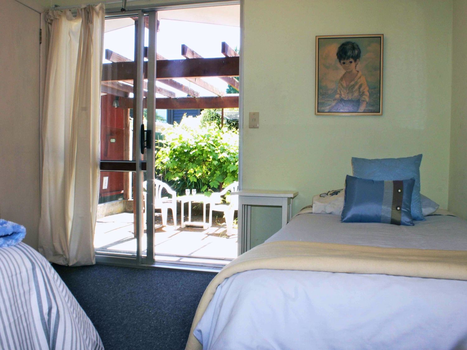 A bedroom with a single bed with blue pillows a side table and sliding glass door leading to an outdoor patio with chairs and a table at Moeroa - Wharewaka Holiday Home in Wharewaka