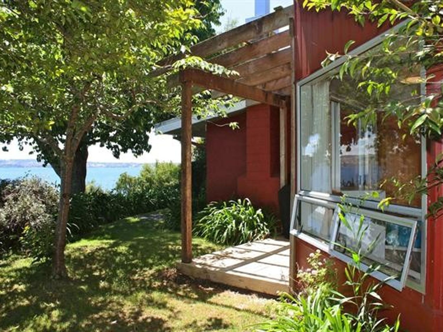 A garden area with trees and plants next to a red house with an open window and a wooden pergola at Moeroa - Wharewaka Holiday Home in Wharewaka