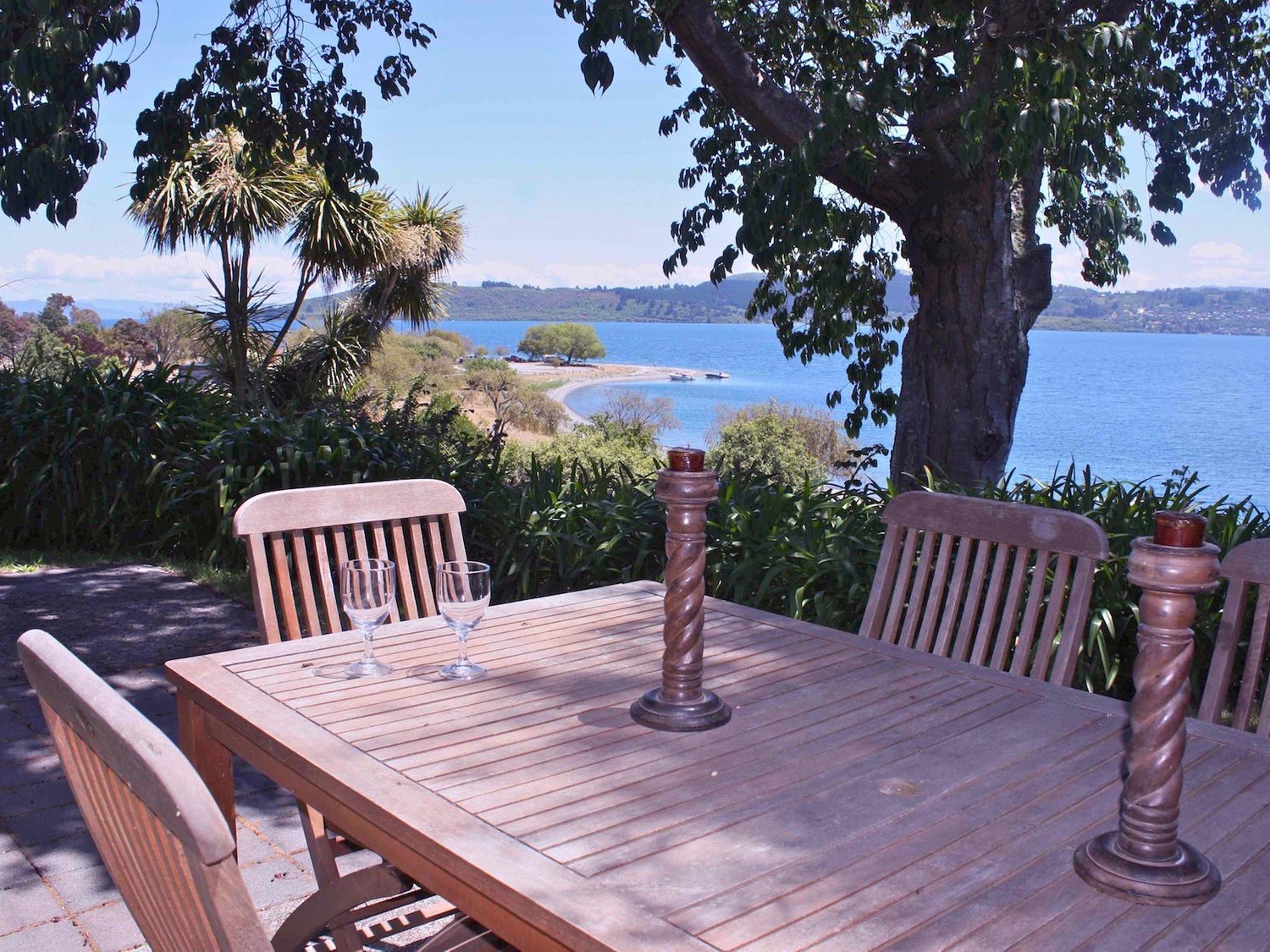 An outdoor wooden table with chairs under trees overlooking a lake and distant land at Moeroa - Wharewaka Holiday Home in Wharewaka