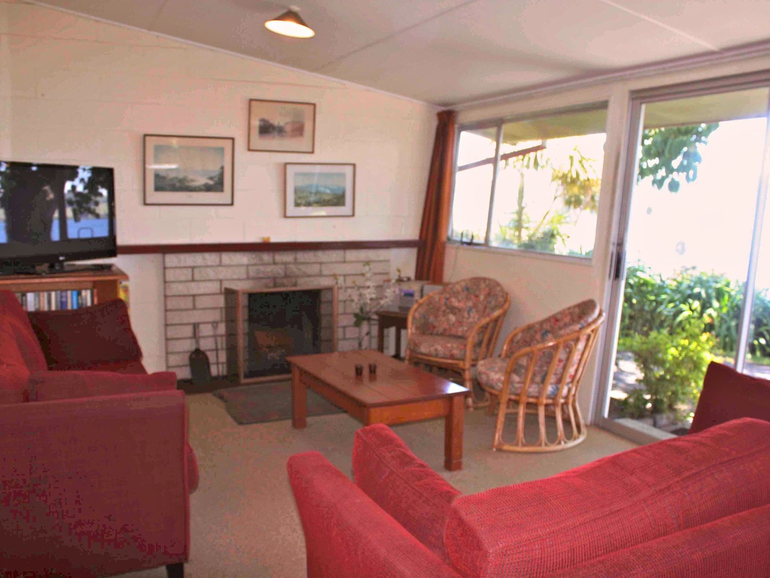A living room with red sofas a wooden coffee table a fireplace and two wicker chairs with floral cushions at Moeroa - Wharewaka Holiday Home in Wharewaka
