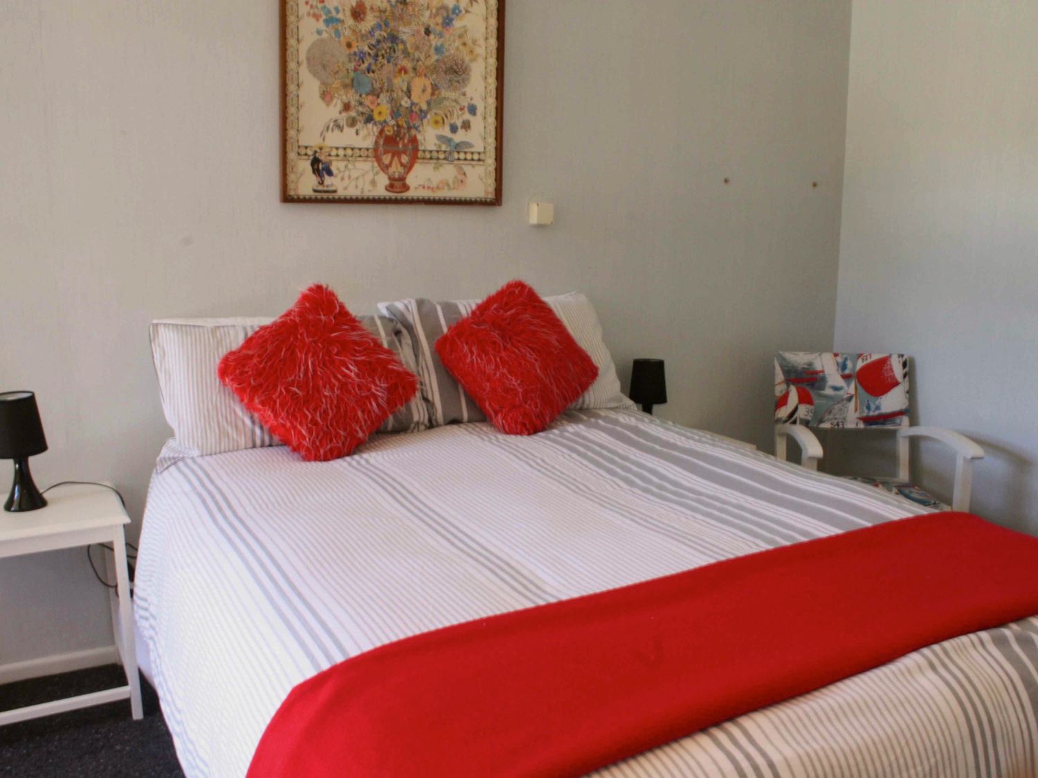 A bedroom with a bed with red cushions and a red throw blanket bedside tables with lamps and a chair with a patterned cushion at Moeroa - Wharewaka Holiday Home in Wharewaka