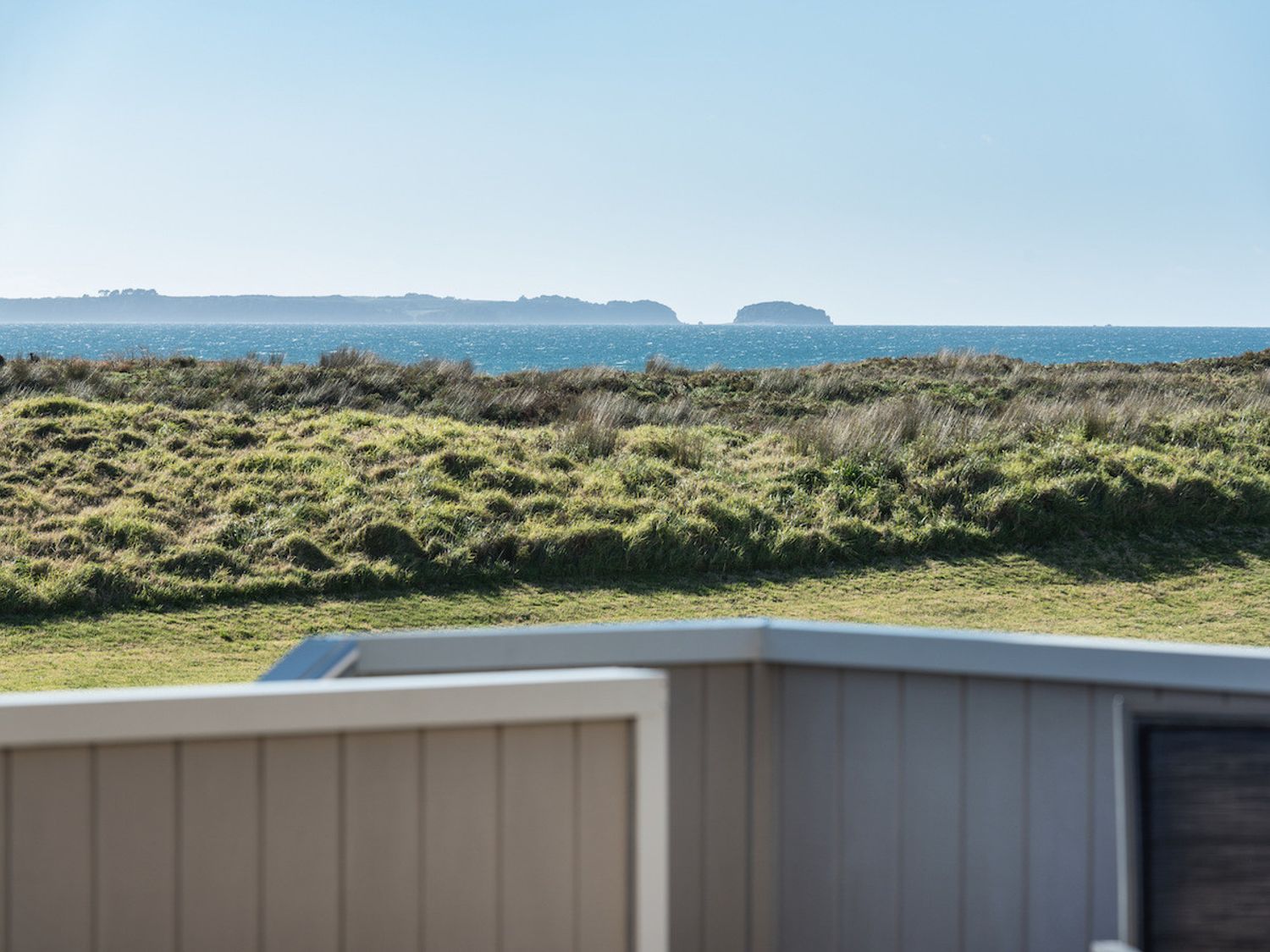 A grassy area with a view of the ocean and distant islands behind a fence at A Beach Barn Haven - Papamoa Holiday Home in Papamoa