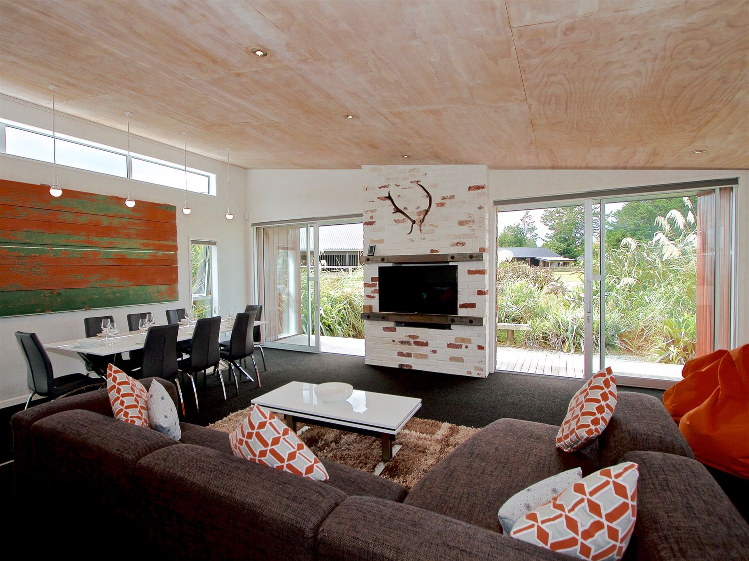 A living room with a brown sectional sofa and patterned pillows a white coffee table a dining table with black chairs and pendant lights a brick fireplace with a mounted tv and large windows with garden views at Stargazer Ohakune Holiday Home Ohakune