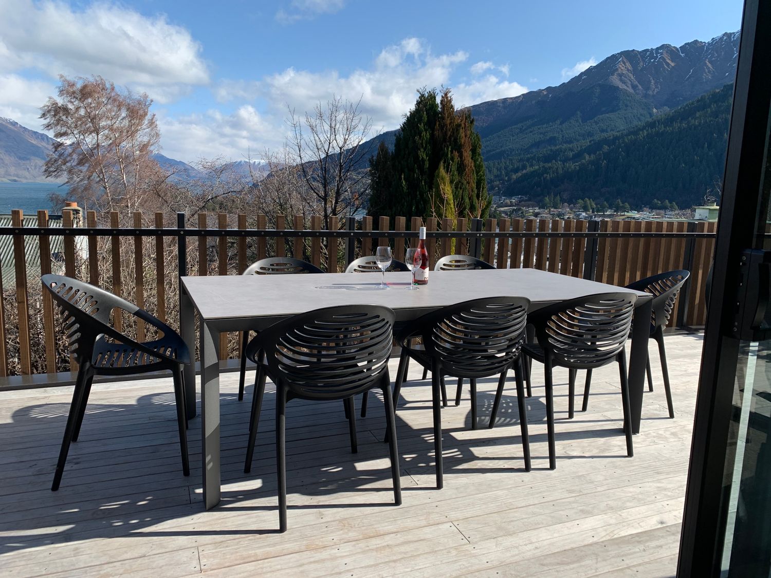 An outdoor deck with a table and eight chairs with wine and glasses and mountains in the background at Central Southern Lakes Queenstown Holiday Home in Queenstown