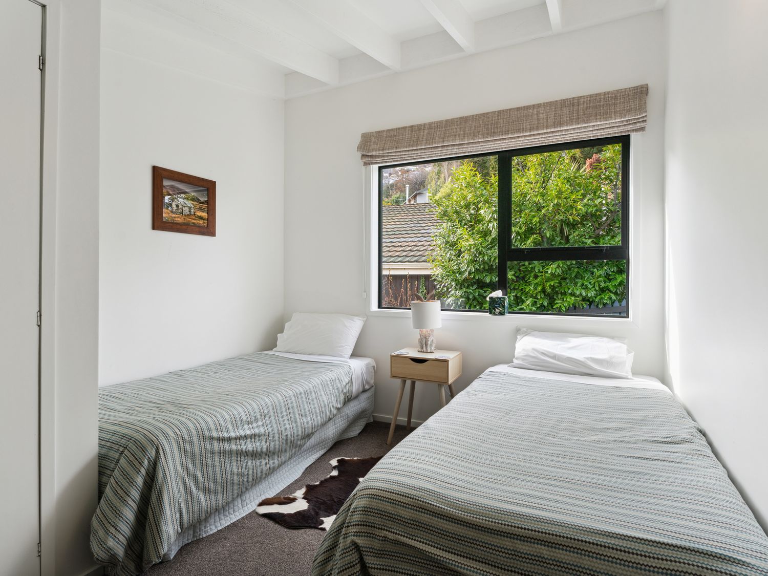 A bedroom with two single beds a nightstand with a lamp and a window showing green trees at Central Southern Lakes - Queenstown Holiday Home in Queenstown