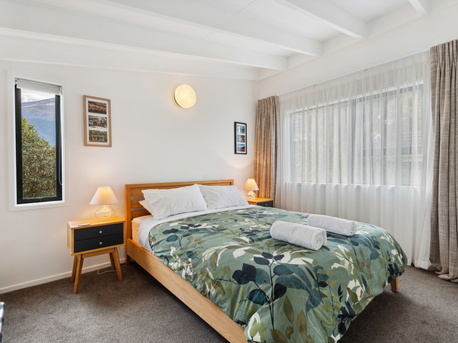 A bedroom with a bed between bedside tables with lamps and windows with curtains at Central Southern Lakes - Queenstown Holiday Home in Queenstown