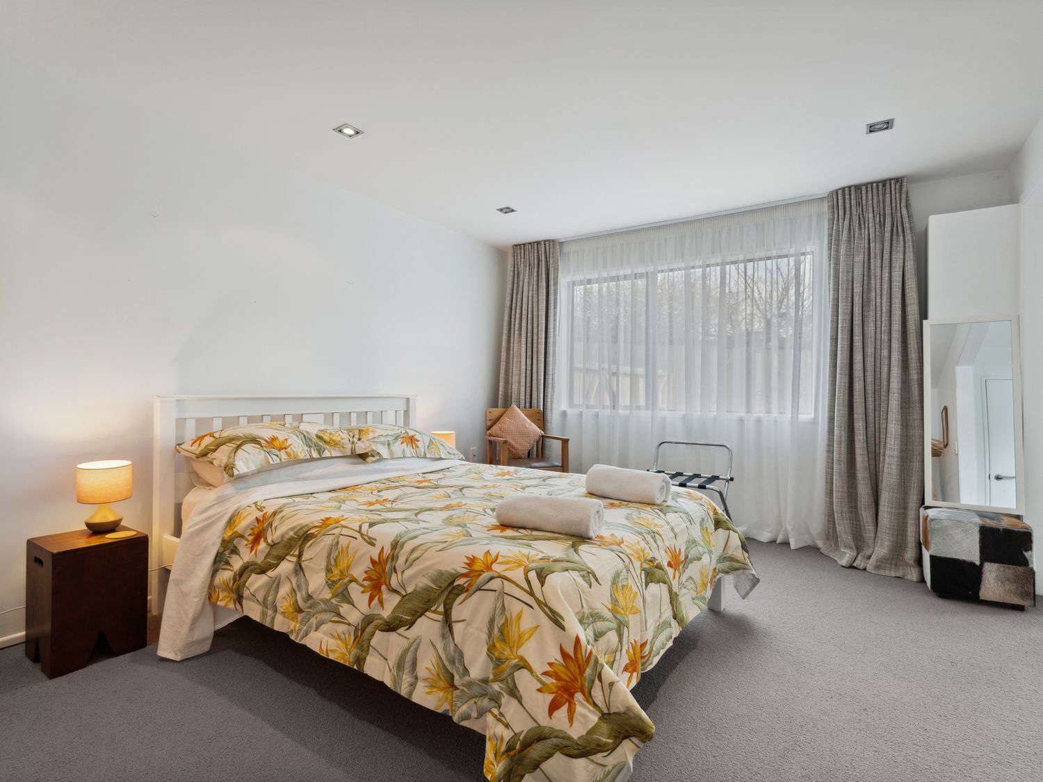 A bedroom with a bed with floral bedding bedside tables with lamps a chair near a window and a standing mirror at Central Southern Lakes - Queenstown Holiday Home Queenstown
