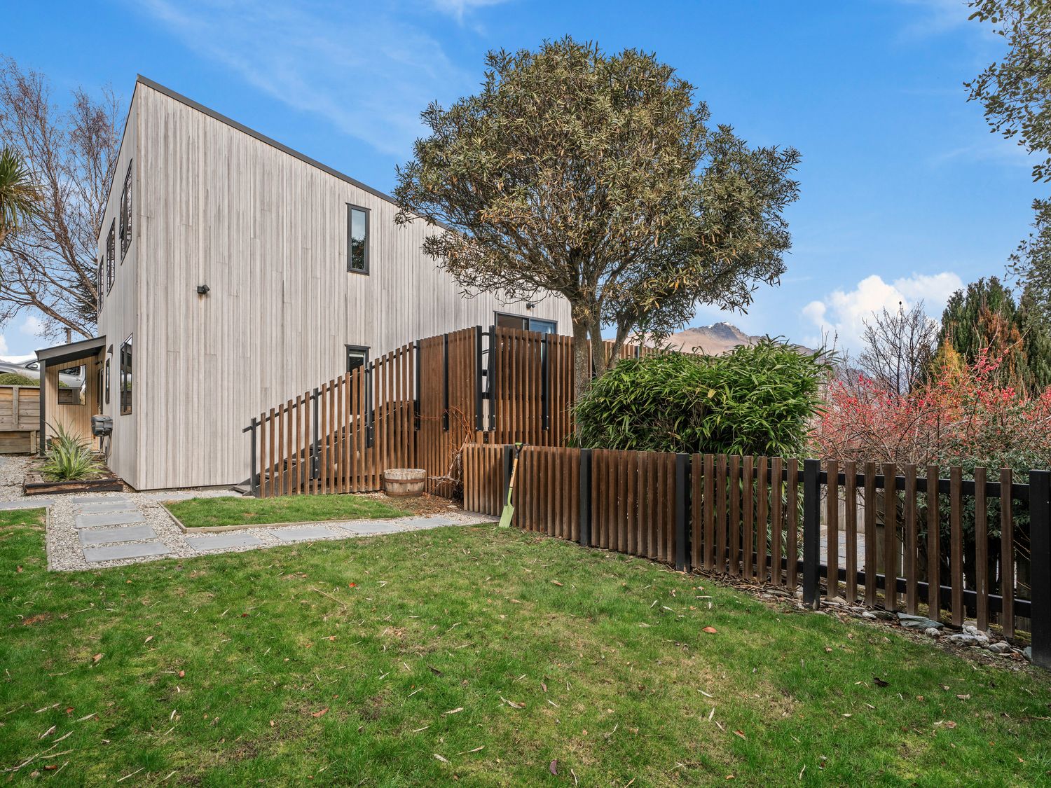 A backyard with grass stone pathway wooden fence and a tree near a modern house at Central Southern Lakes Queenstown Holiday Home in Queenstown