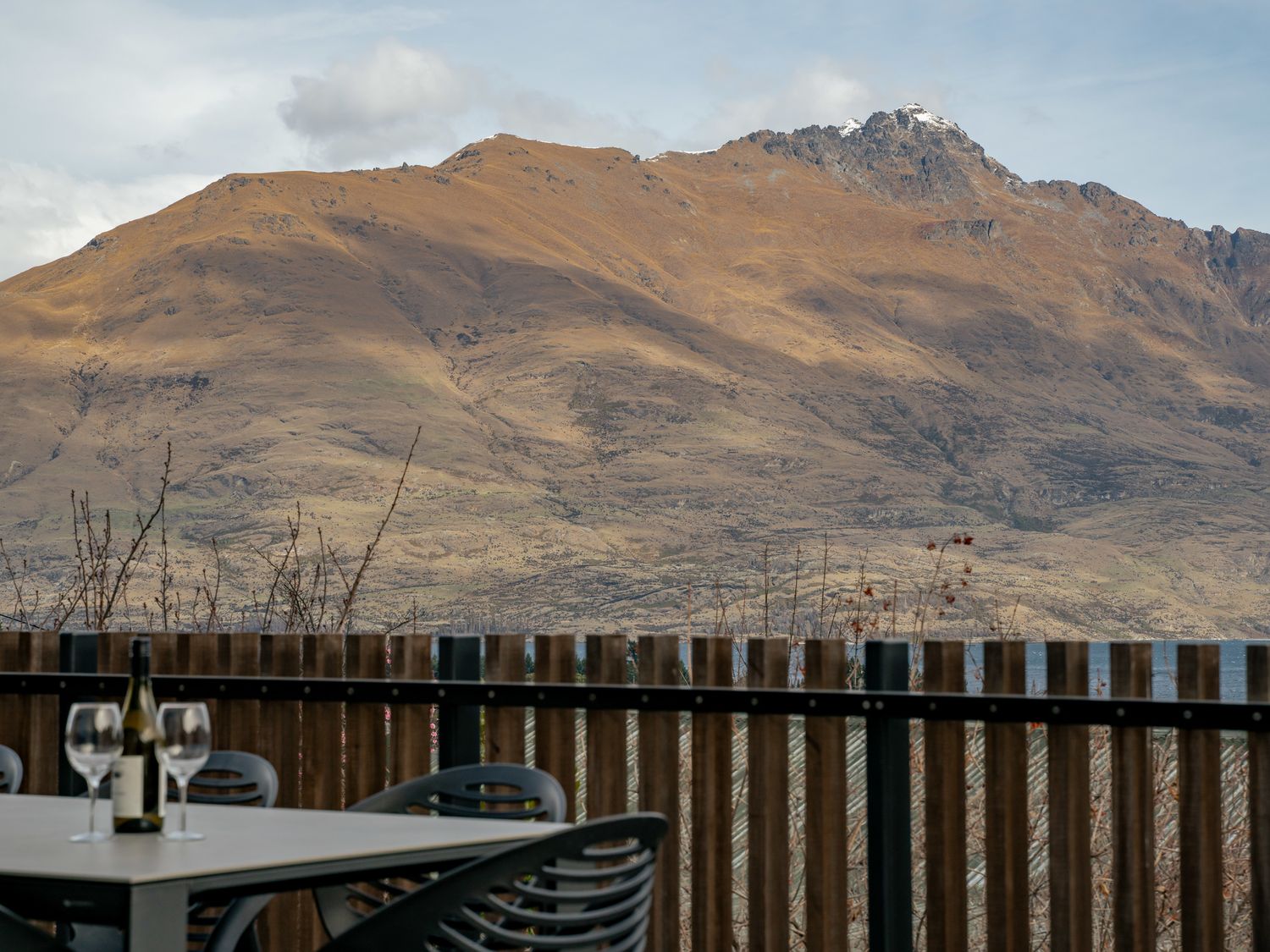An outdoor table with a bottle and two wine glasses on a balcony with a wooden fence and a mountain in the background at Central Southern Lakes Queenstown Holiday Home Queenstown