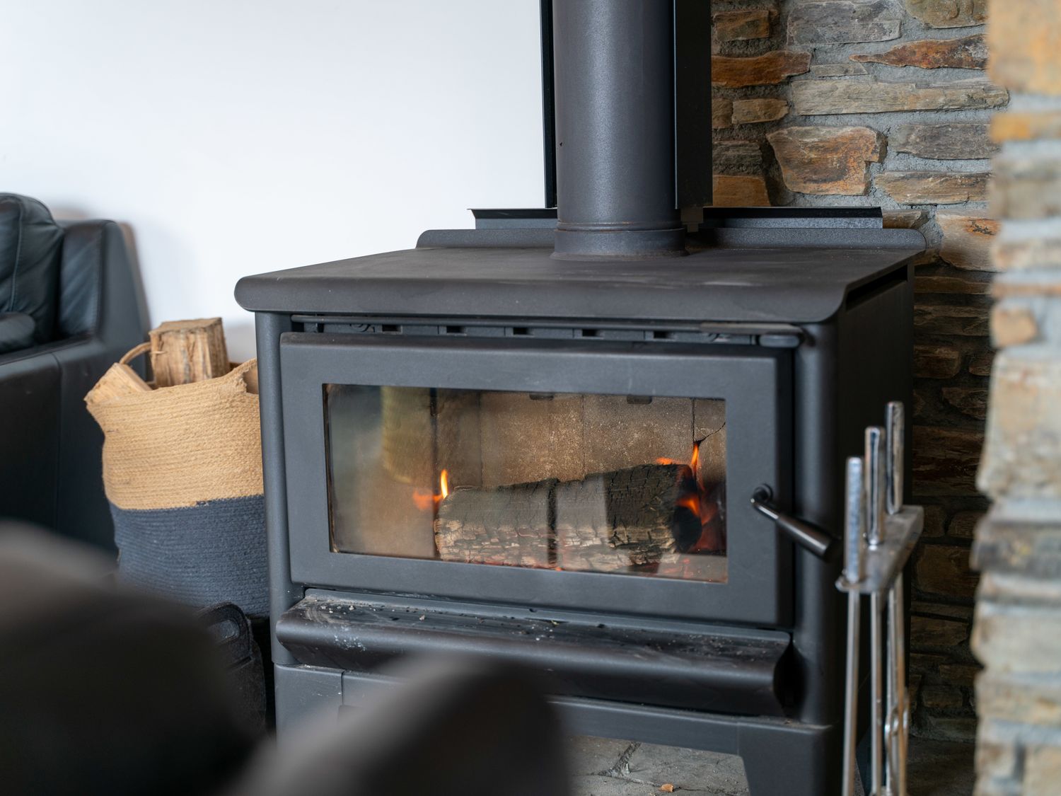 A wood-burning stove with firewood inside a room with a stone wall and a basket of firewood at Central Southern Lakes - Queenstown Holiday Home in Queenstown