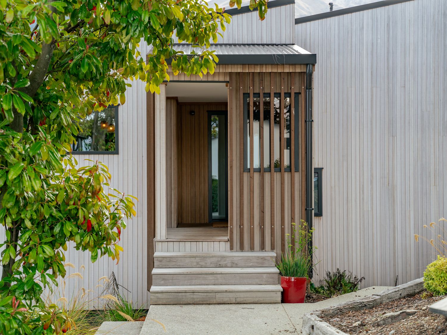 A house entrance with wooden steps and vertical wood paneling surrounded by plants and trees at Central Southern Lakes - Queenstown Holiday Home in Queenstown